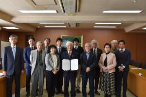 第12回市民憲章市民会議にて撮影された石川良三市長や出席者による記念写真。市長は春日部市民憲章（案）を手にしてこちらに向けて広げている。