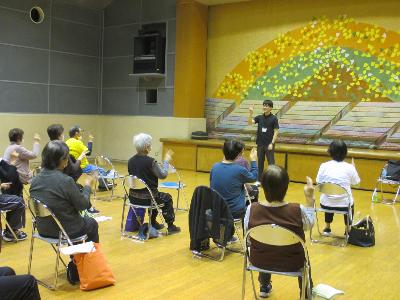 講師と参加者が関節痛や体力低下予防の体操を行っている様子の写真