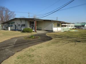 雲一つない晴天の下建つ茶色の瓦屋根が特徴的な春日部市立第6保育所の外観を目前に広がる芝生側から撮影した写真