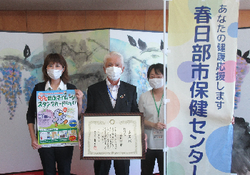 屏風の前で、表彰状を持った男性と、ポスターとのぼりをそれぞれ持った女性たちが並んでいる写真