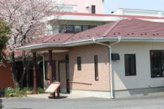 赤い屋根のある建物と、その横に桜の木が立っている、春日部市第2地域包括支援センターの外観の写真