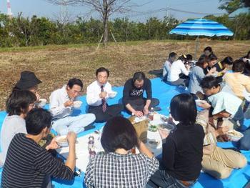 工場内の緑地帯にシートを広げ大勢で芋煮をほおばる社員の写真