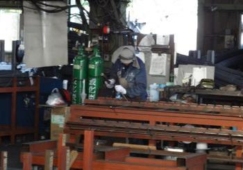 群青色の作業服に灰色のエプロン、溶接帽子 さらに白い軍手をし作業台の上で作業をしている人の写真