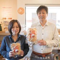 店舗内でオーナーの工藤ご夫妻が胸の前に商品化されたおせんべいを持っている正面写真