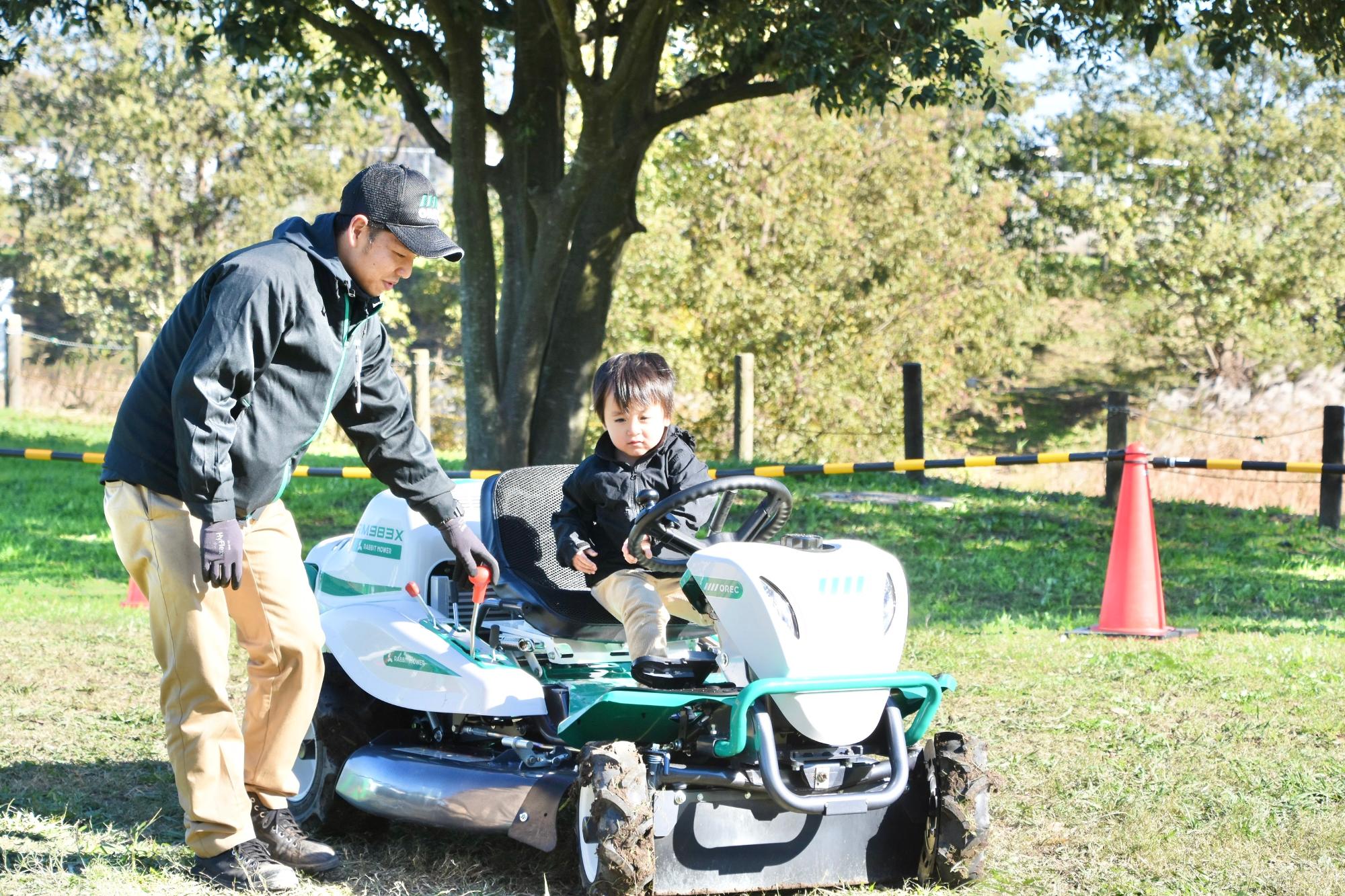 草刈り機に乗るこどもと補助するスタッフ