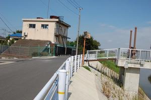 晴れた空の下、川にかかる橋の横に緑色のフェンスで囲まれて建っている藤塚ポンプ場の外観の写真