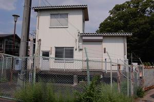 晴れた空の下、大きな1本の木と緑色の網目状のフェンスに囲まれるようにして建っている旧倉松川排水機場の外観の写真