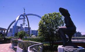 雲一つない晴天の下そびえ立つ古利根公園橋の麦わら帽子を模したモニュメントとその前方にあるブロンズ像の写真