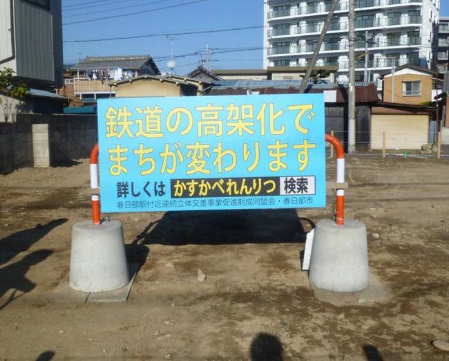 春日部駅付近の住宅地にある市の土地の地面に設置された水色のPR看板には黄色い文字で「鉄道の高架化でまちが変わります」と書かれている状態の写真