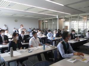 マイクを持ち立って質疑応答する男性と机に座り耳を傾ける多数の参加者の写真