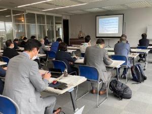 ミーティングで机に座りスクリーンに目を向ける参加者たちの写真