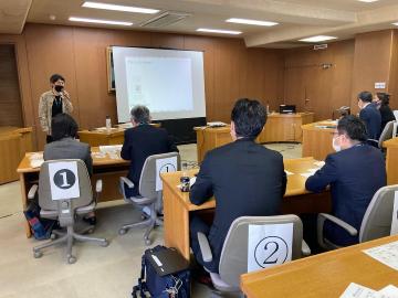 スクリーンの横に立ち話をする男性と真剣に聞き入る参加者の様子の写真