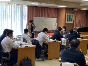 ホワイトボードに向かい説明をするファシリテーターの話を聞く参加者の写真