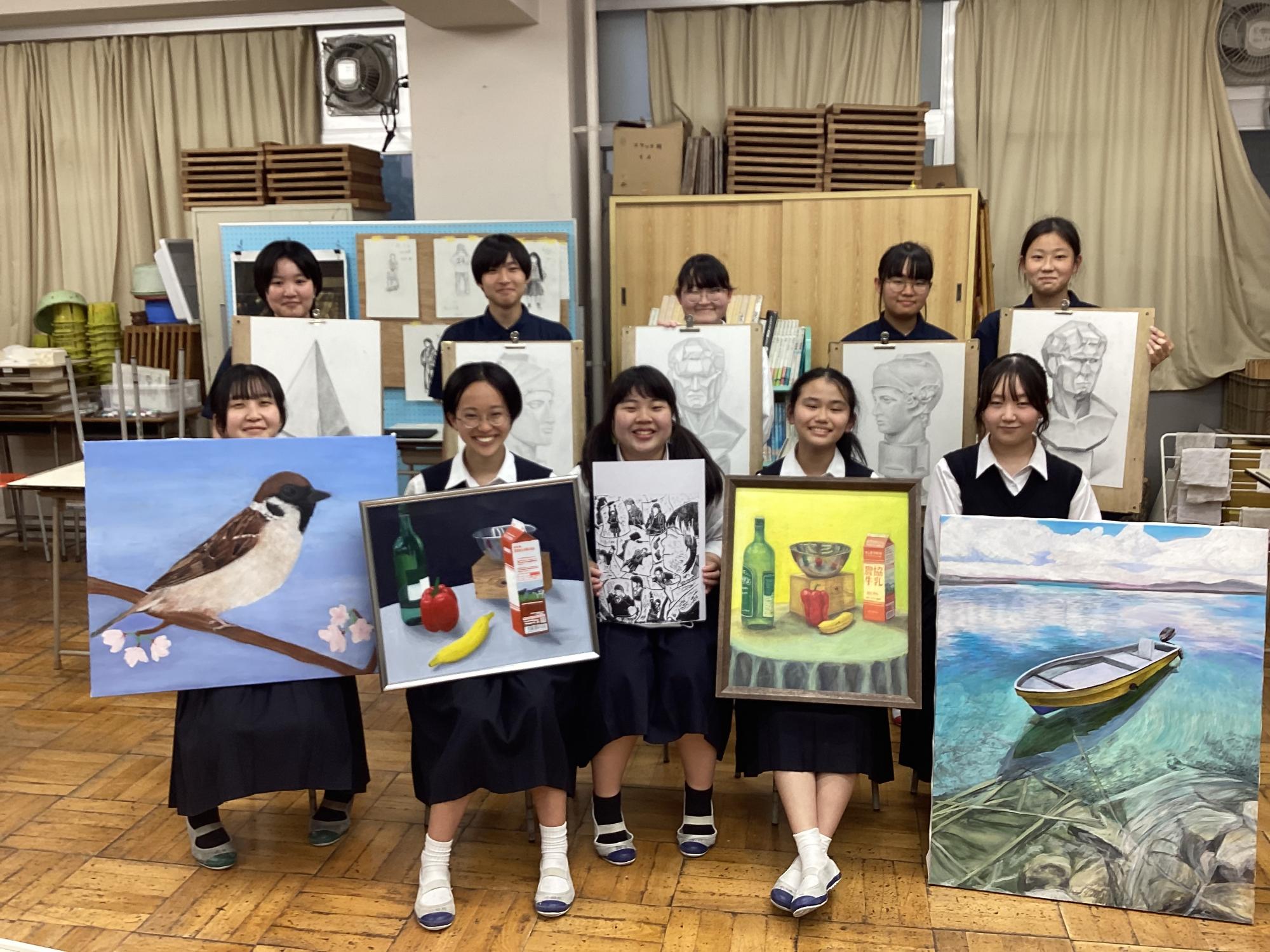 10名の学生が美術作品を手に持ち集合写真を撮る様子