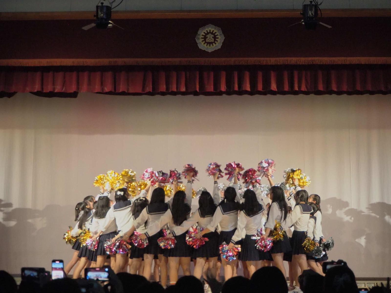 女子高校生10名くらいが後ろ向きでステージでポーズをとる様子