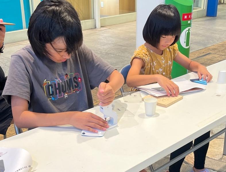 キリで穴を開けた店舗バナーに、機械を使ってボタンを留める女子児童の様子