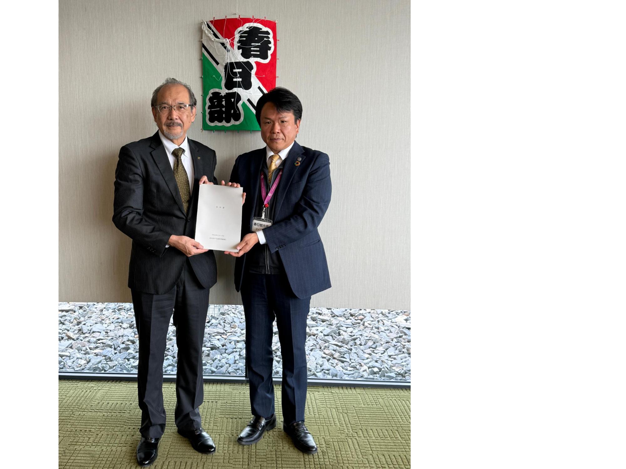 春日部と書かれた凧の前で、審議会会長から答申を受け取る岩谷市長の写真