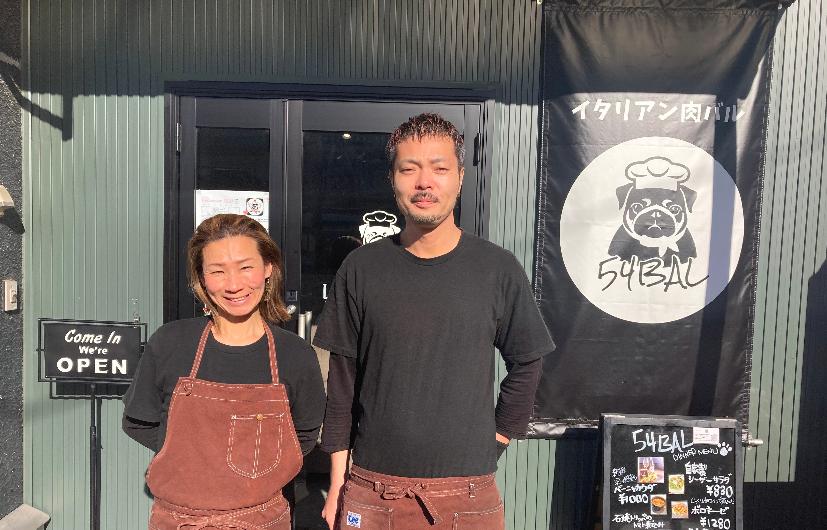 お店のオリジナルTシャツとえんじ色のエプロンを身に着けてお店の前で微笑む五嶌弘太郎さん(右)と真理さん(左)