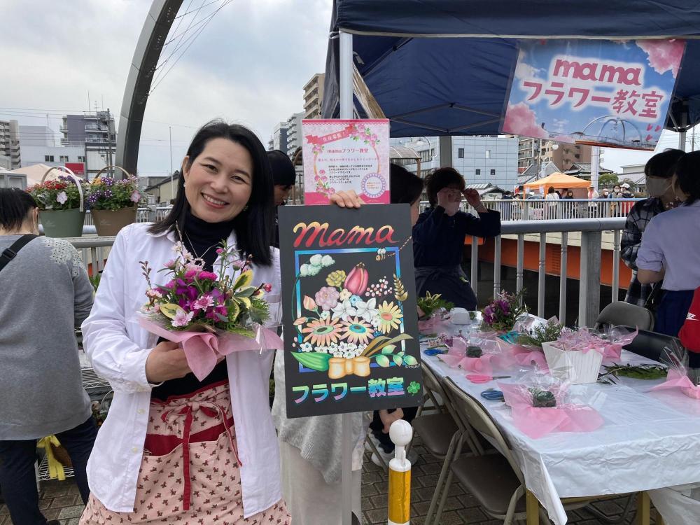 フラワーアレンジメントと看板を手に笑うmamaフラワー教室の白石さん