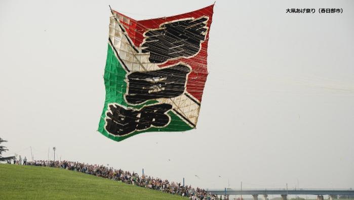 「春日部」と大きく書かれた赤・緑・白の3色カラーの大凧が空に上がっている写真