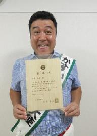 桐製の委嘱状を持ってカメラのほうへ向ける山崎さんの写真