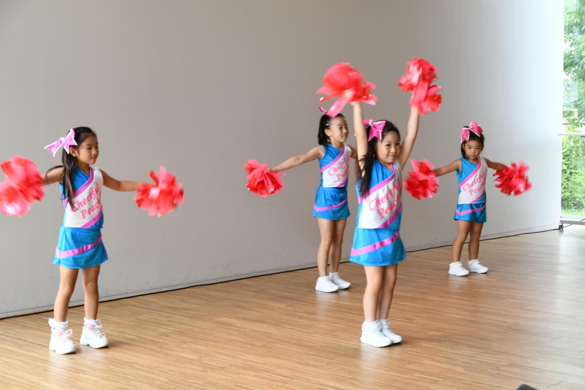 チーム名の書かれた水色とピンクのお揃いの衣装を着て赤いポンポンを持ち踊る4人の女の子たちの写真