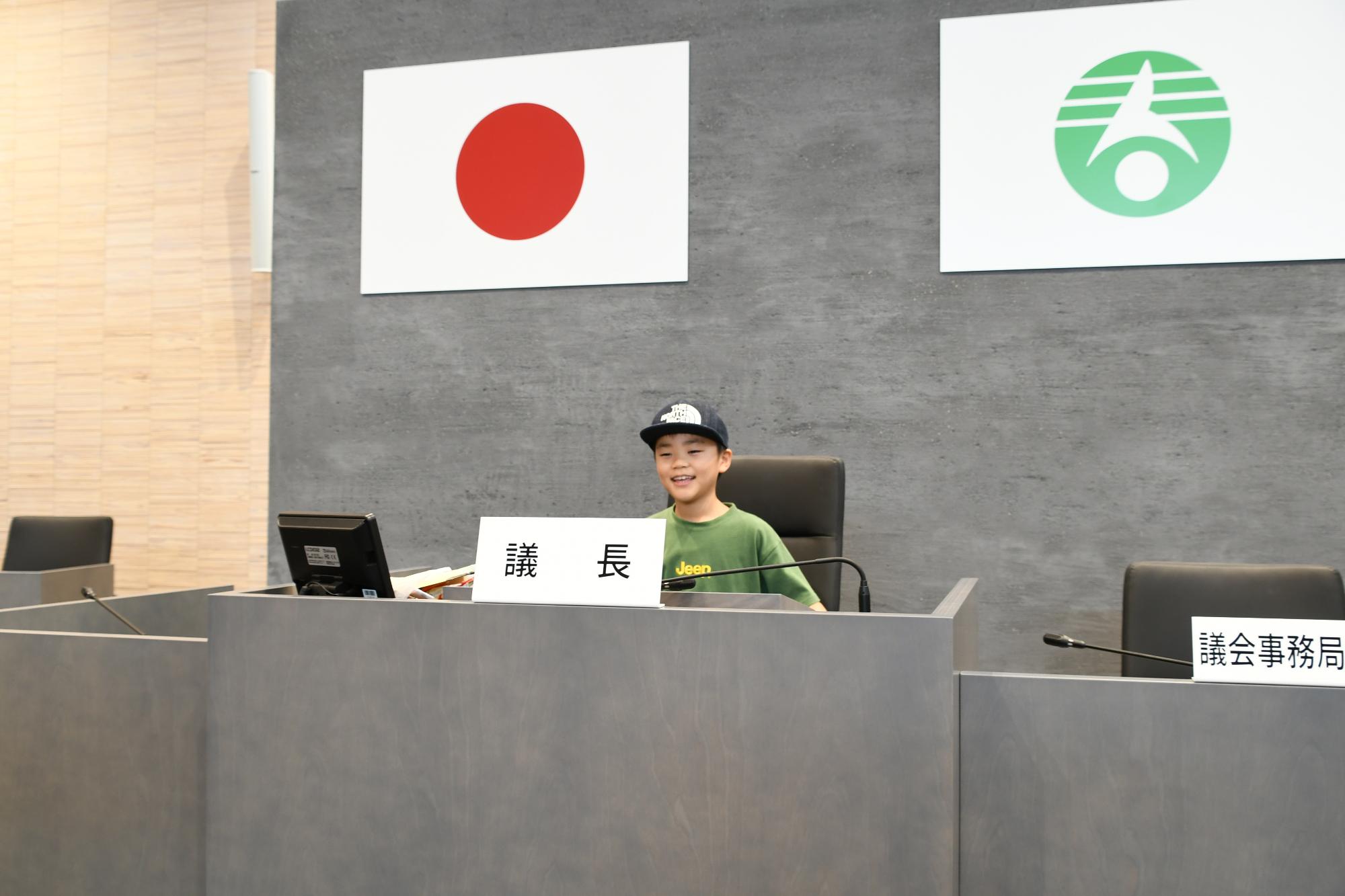 議長席に座る男の子の写真。黒色の防止をかぶり、深緑色のTシャツを着て、笑顔を浮かべている。