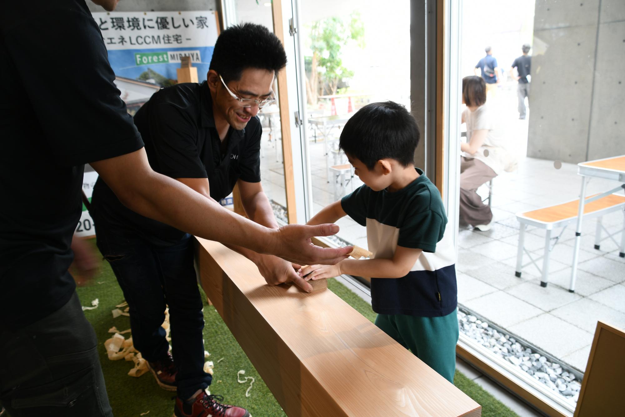 緑色と白色と紺色のストライプ柄のTシャツと緑色のズボンを着た男の子がカンナ削りを体験している様子。