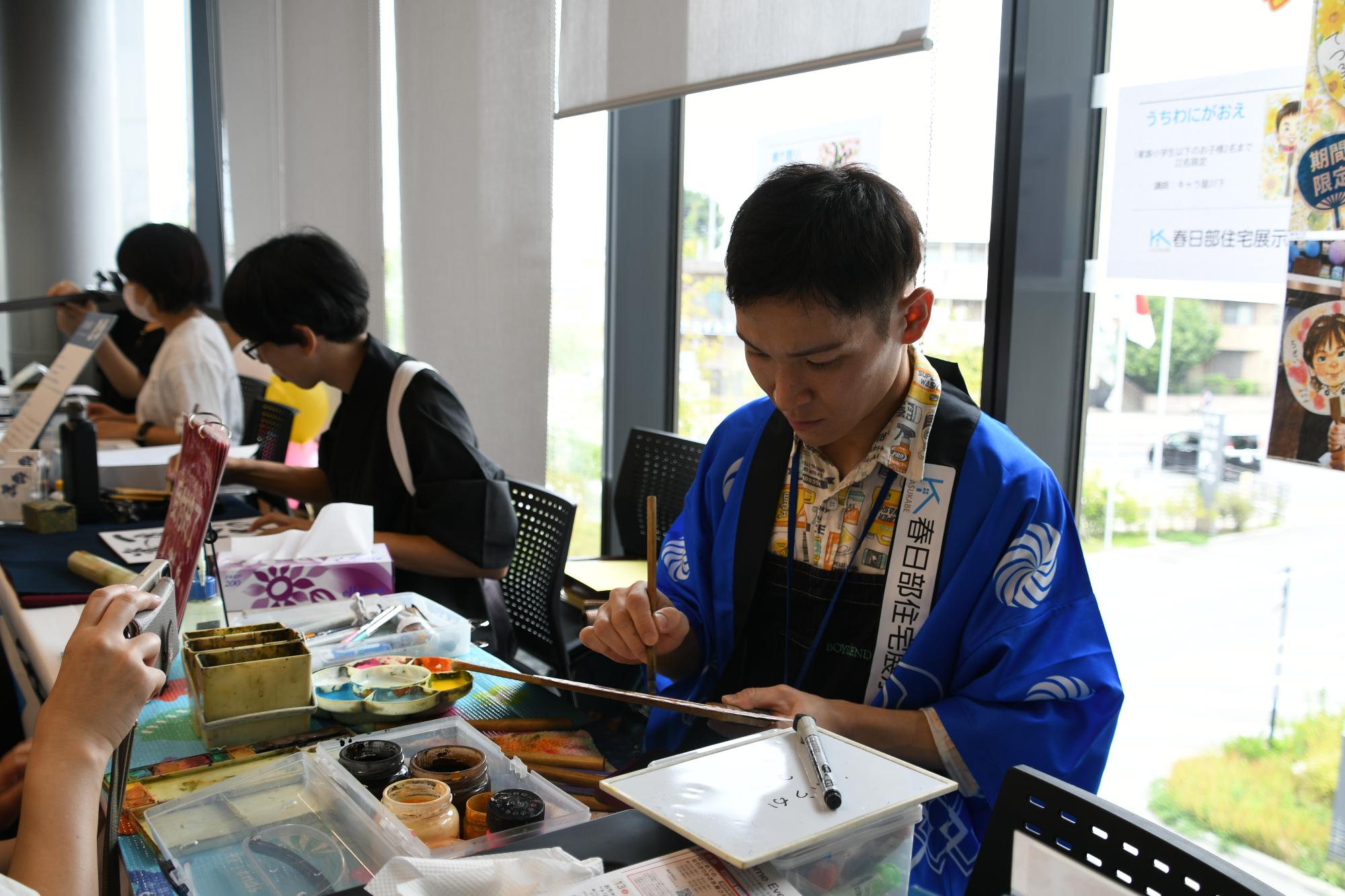 「春日部住宅展示場」と書かれた青いはっぴを着て参加者の似顔絵を描く男性と黒色の着物で名前書きを行う男性が写った写真