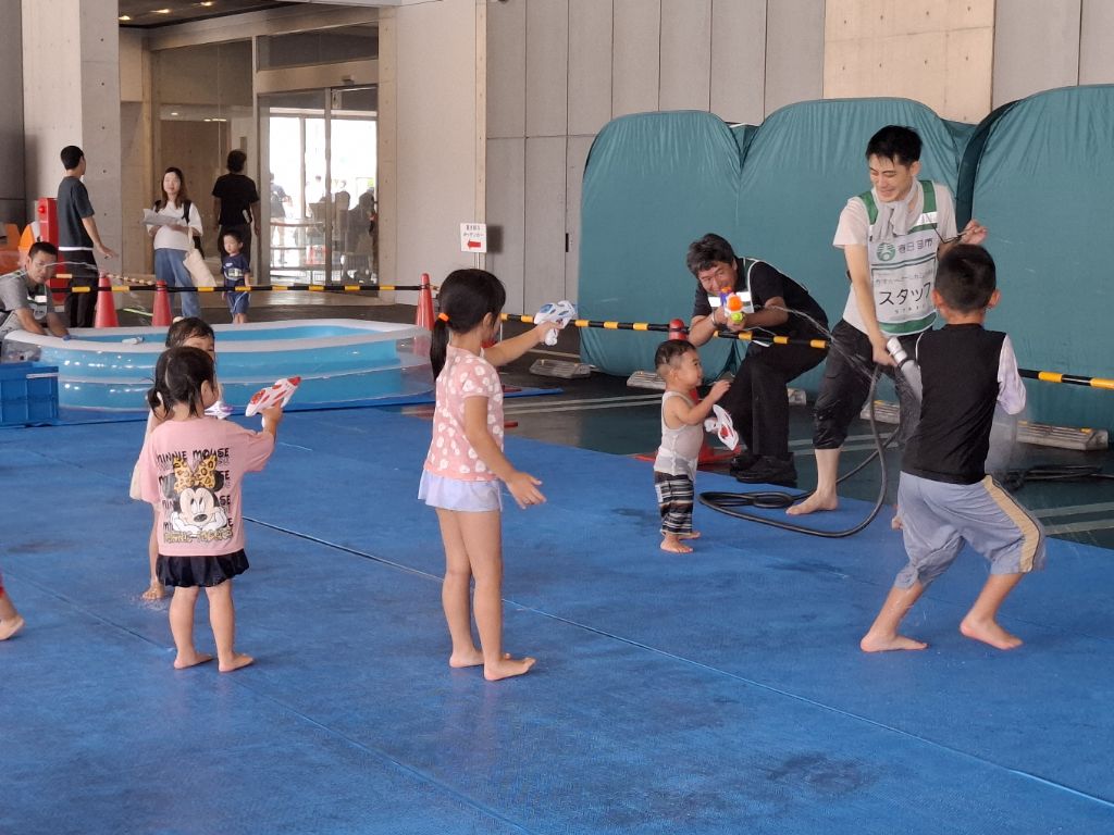 「水遊びdeクール大作戦」に参加しているこどもたちとシャワーや水鉄砲で放水するスタッフの写真