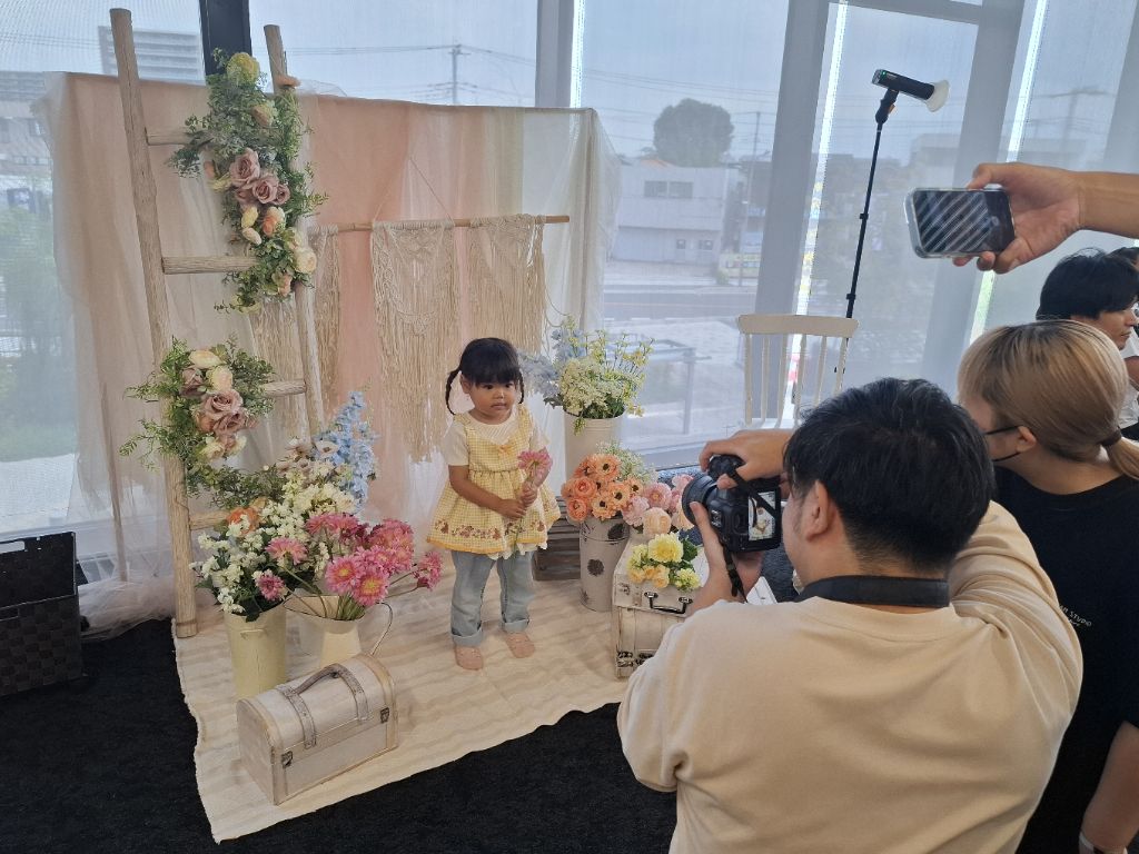 カラフルな花に囲まれた撮影スペースでピンクをお花を手に持って写真に写る女の子とカメラをのぞき込むスタッフの写真