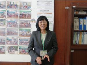 写真や資料の貼ってある壁を背景に、笑顔でインタビューに応じる折原栄養教諭の写真