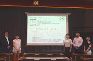 暗幕のかかった部屋でスクリーンにスライドが映し出され、5名の男女がその脇に立っている写真