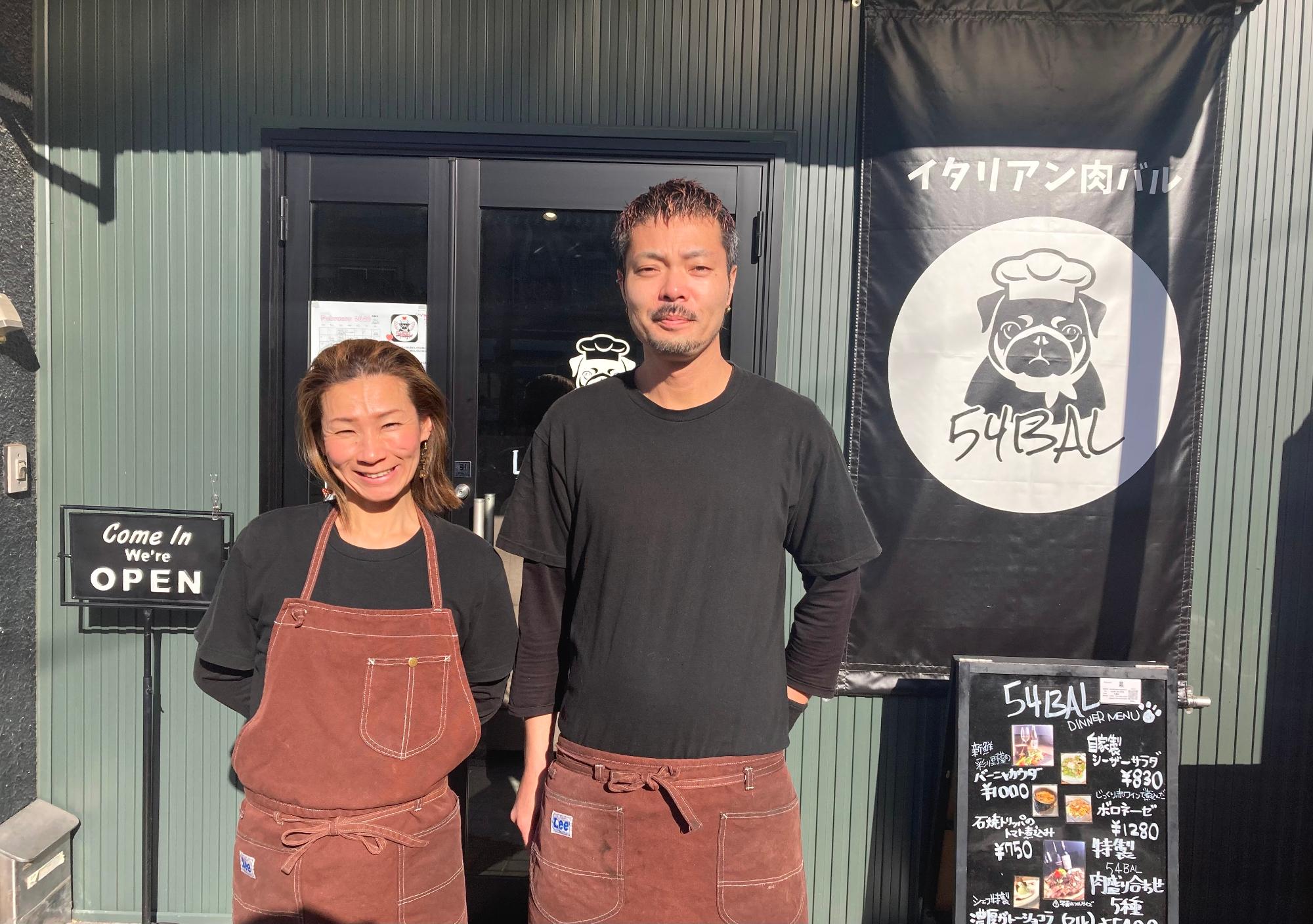 黒いTシャツとえんじ色のエプロンを着て、お店の前で微笑む五嶌弘太郎さん、真理さん