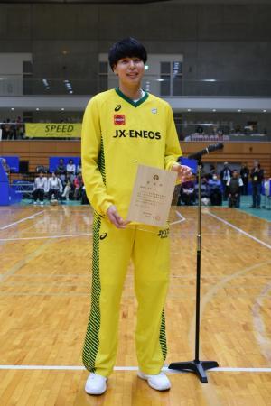 体育館で委嘱状を持ち笑顔を見せるユニフォーム姿の渡嘉敷さんの写真