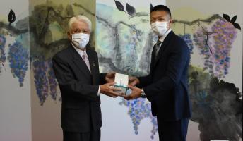 藤が描かれた屏風の前で市長にマスクを手渡ししている内山さんの写真