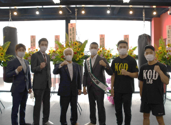 オープン祝いの花を背景にスーツ姿の男性6名が並んで記念撮影している写真