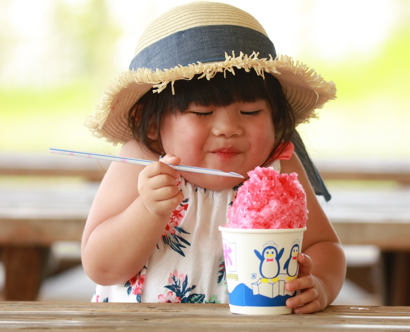 イチゴ味のかき氷をおいしそうに食べる麦わら帽子をかぶり、花柄のワンピースを着た女の子