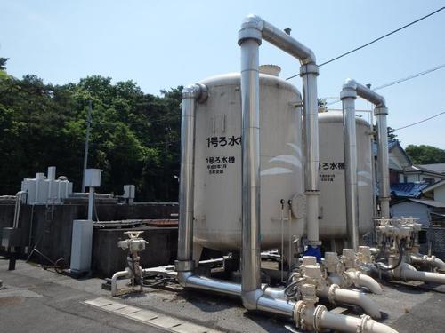 青空の下木々に囲まれて設置された配水管が接続されたろ水機と印字された巨大タンクの写真