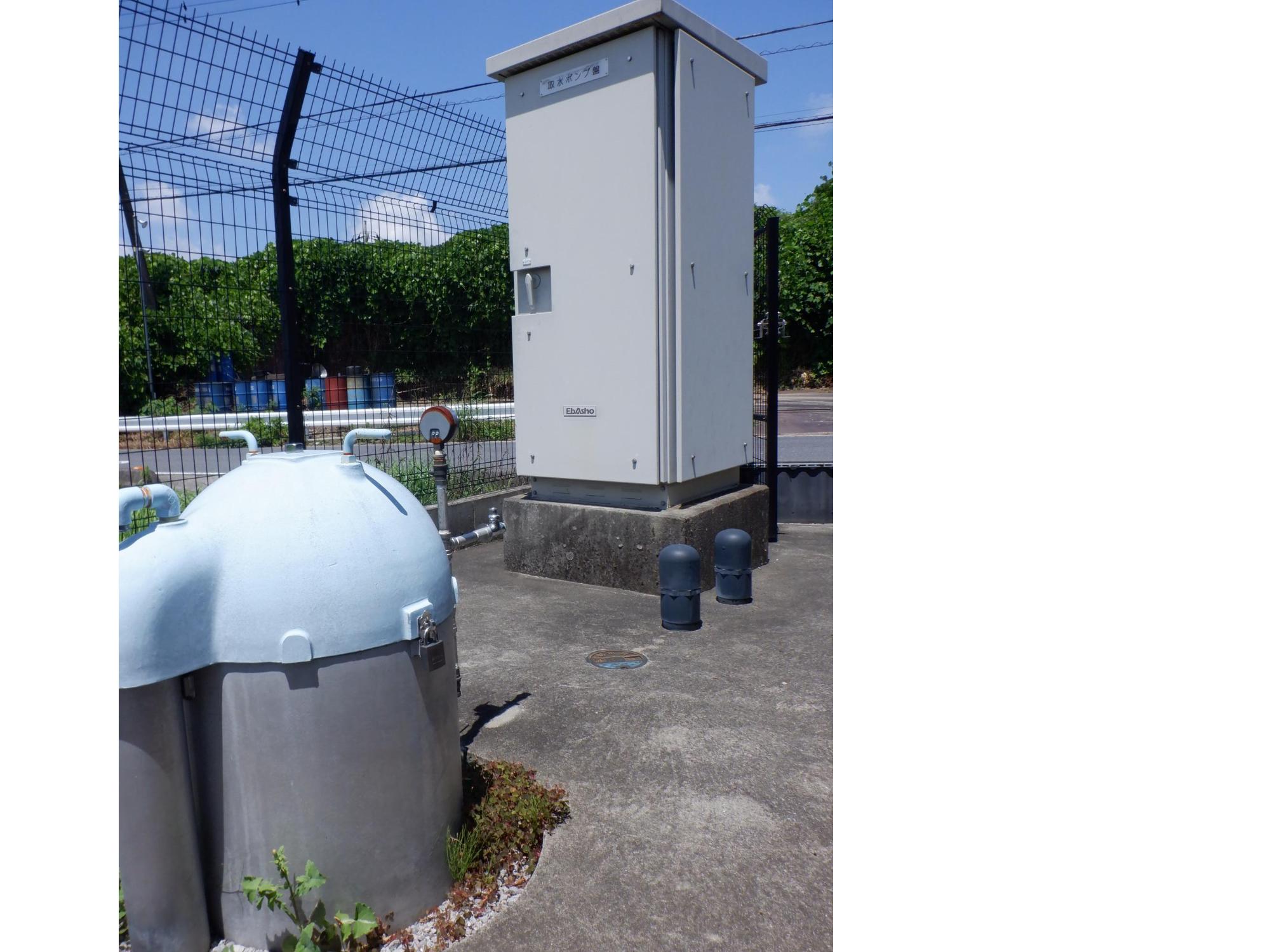 コンクリートの基礎上に建てられた深井戸とその右横に間隔を空けて設置されたポンプ制御盤の写真