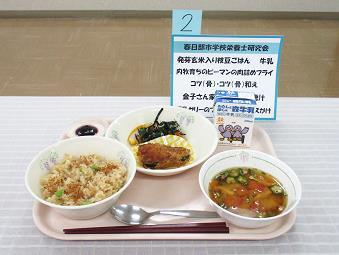 お盆の上にお椀に盛られた発芽玄米入り枝豆ごはんとトマトの汁みそ汁を並べピーマンの肉詰めフライやブルーベリーソースのかかった豆乳ゼリーを添えた給食の写真