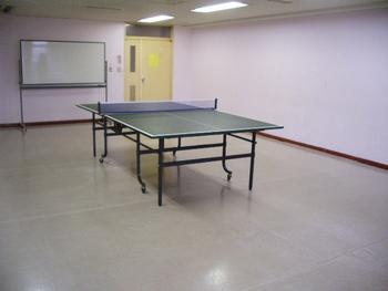 ピンク色の壁と木製フローリングの床の豊春第二公民館第二会議室（卓球台を置いた状態）の写真
